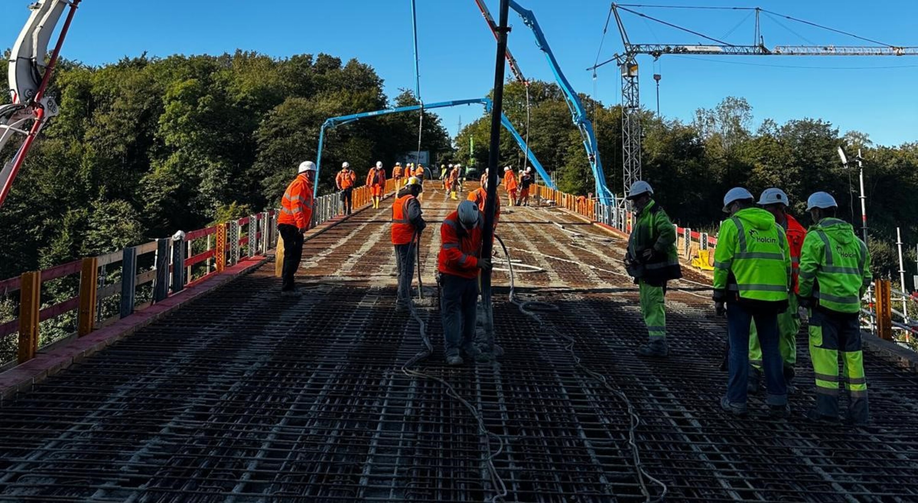 Bauarbeiter in orangefarbener und gelber Schutzkleidung arbeiten auf einer Brücke, deren Oberfläche aus einer dichten Stahlbewehrung besteht. Mehrere Betonpumpen mit langen Auslegern sind im Einsatz und verteilen frischen Beton. Die Szene ist von oben fotografiert, im Hintergrund sind Bäume und blauer Himmel zu sehen.

                                    